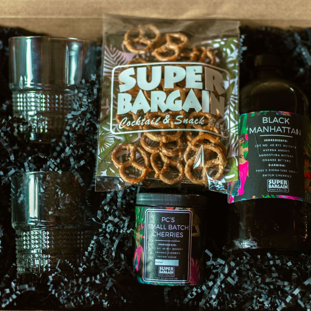 A gift box contains cocktail-themed items, including two textured glass tumblers, a bag of pretzels, a jar labeled "PC's Small Batch Cherries," and a bottle of "Black Manhattan" or "PB&J Old Fashioned" Premium Spirit RTD Cocktail mix with visible ingredients. Black crinkle paper cushions the items. This exclusive set is brought to you by Super Bargain Shop.