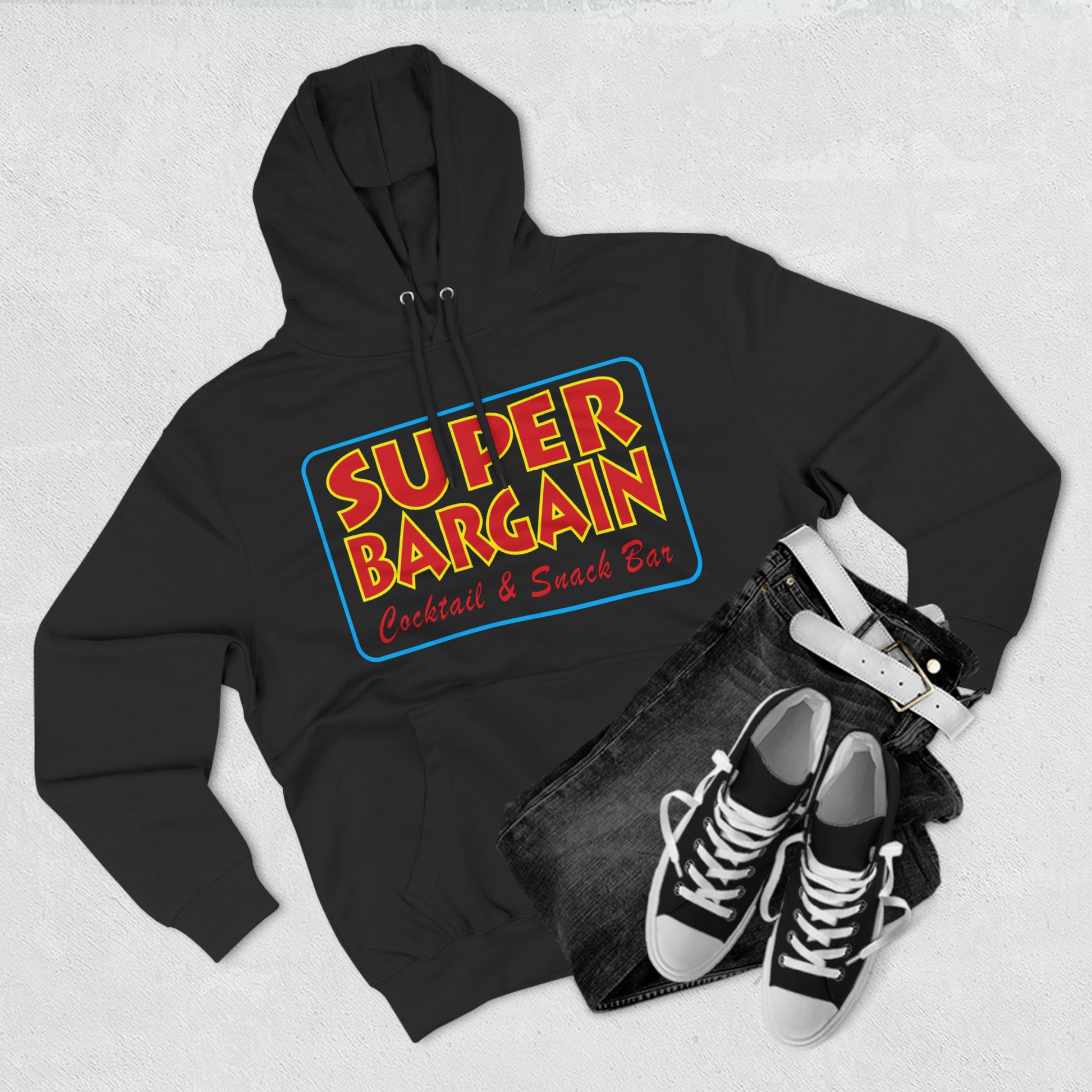 A black Unisex Premium Pullover Hoodie with "Cabbagetown, Toronto Super Bargain Cocktail & Snack Bar" logo, paired with jeans and sneakers, laid out on a textured white background.