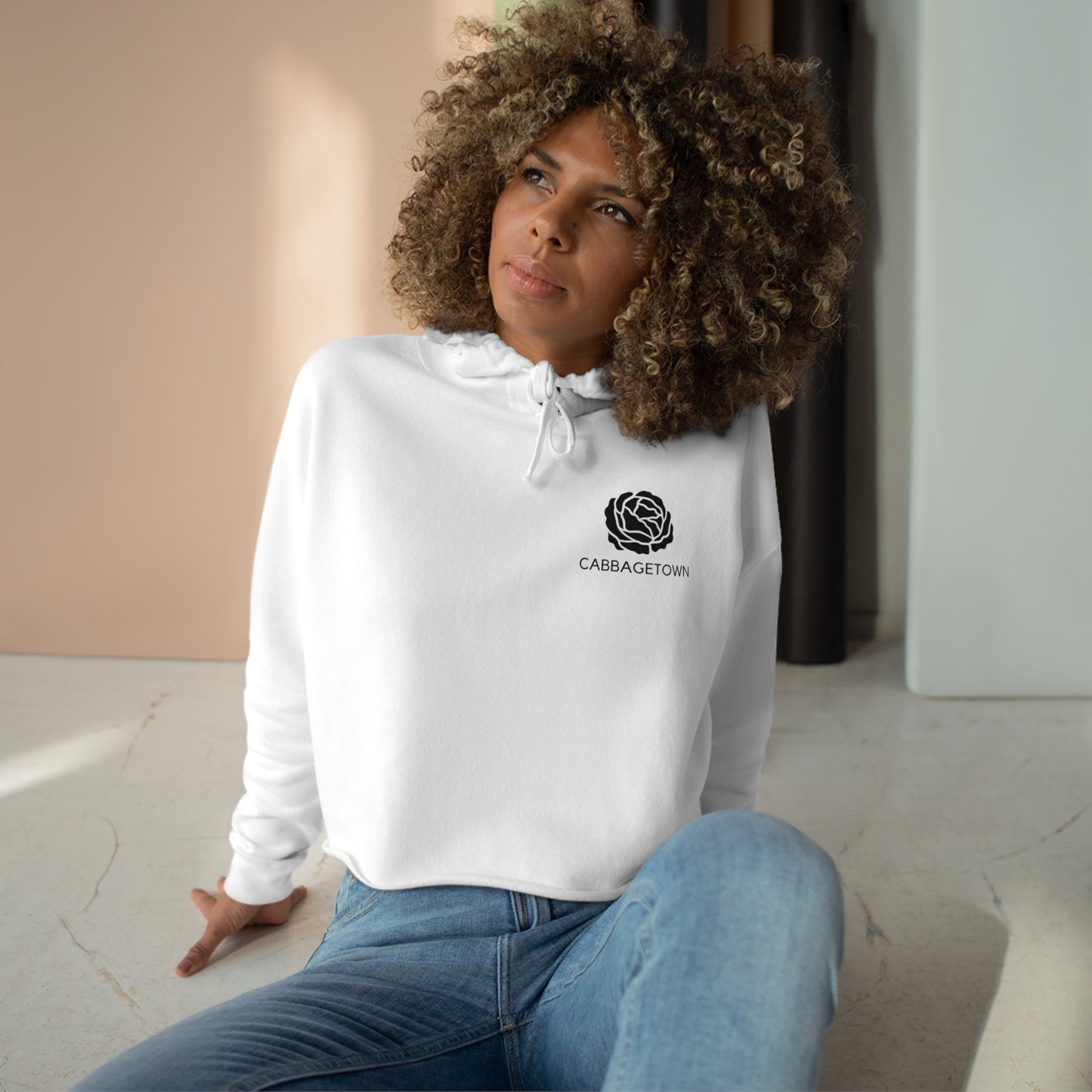 A woman with curly hair in a white Crop Hoodie with Monochrome Cabbagetown and Super Bargain Logo and jeans sits on the floor of a Cabbagetown apartment in Toronto, looking contemplative, with sunlight casting shadows nearby. (Brand Name: Printify)