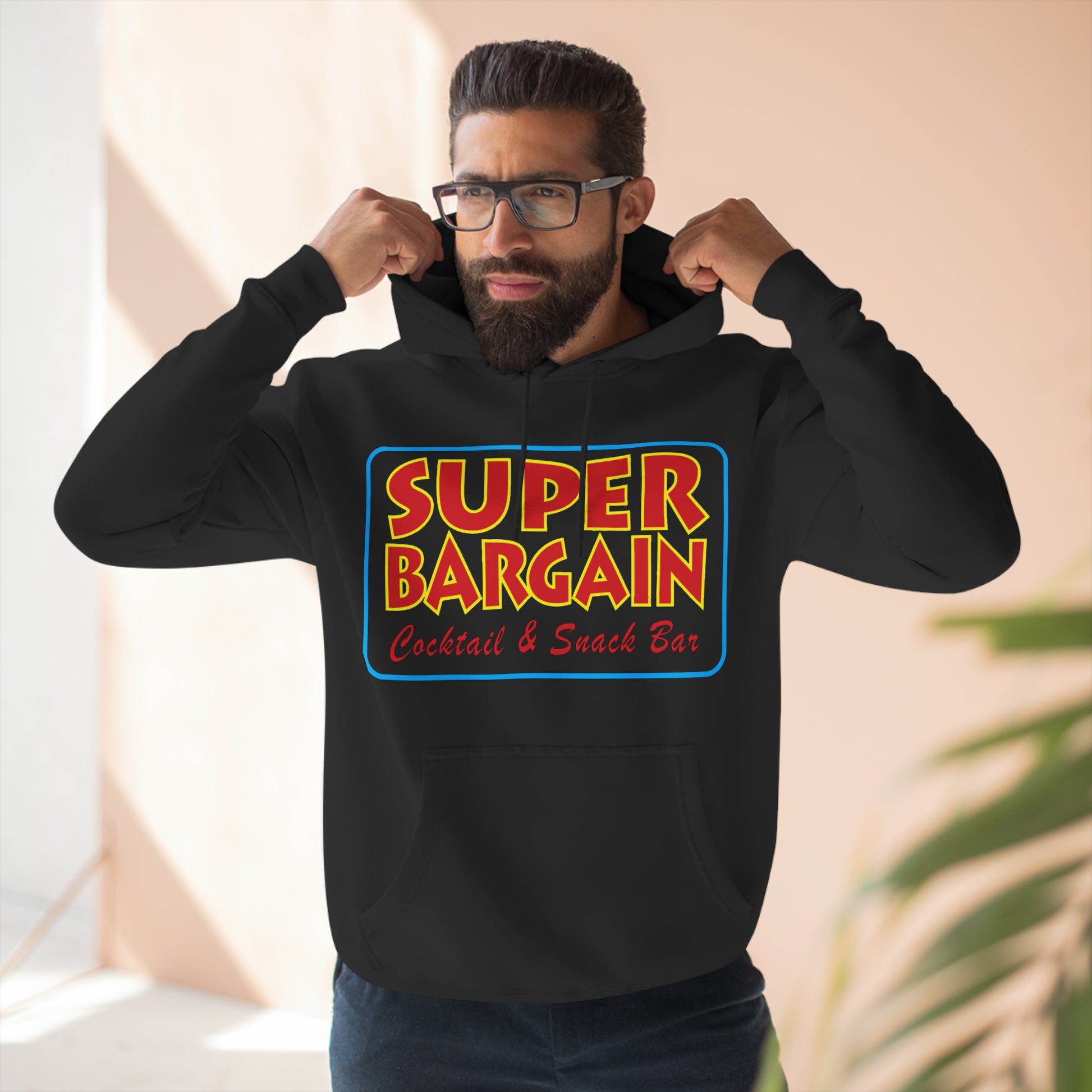 A bearded man in glasses adjusting his black Unisex Premium Pullover Hoodie from Printify, featuring a colorful "Super Bargain Cocktail & Snack Bar" logo on the front, standing against a soft-focus Cabbagetown background.