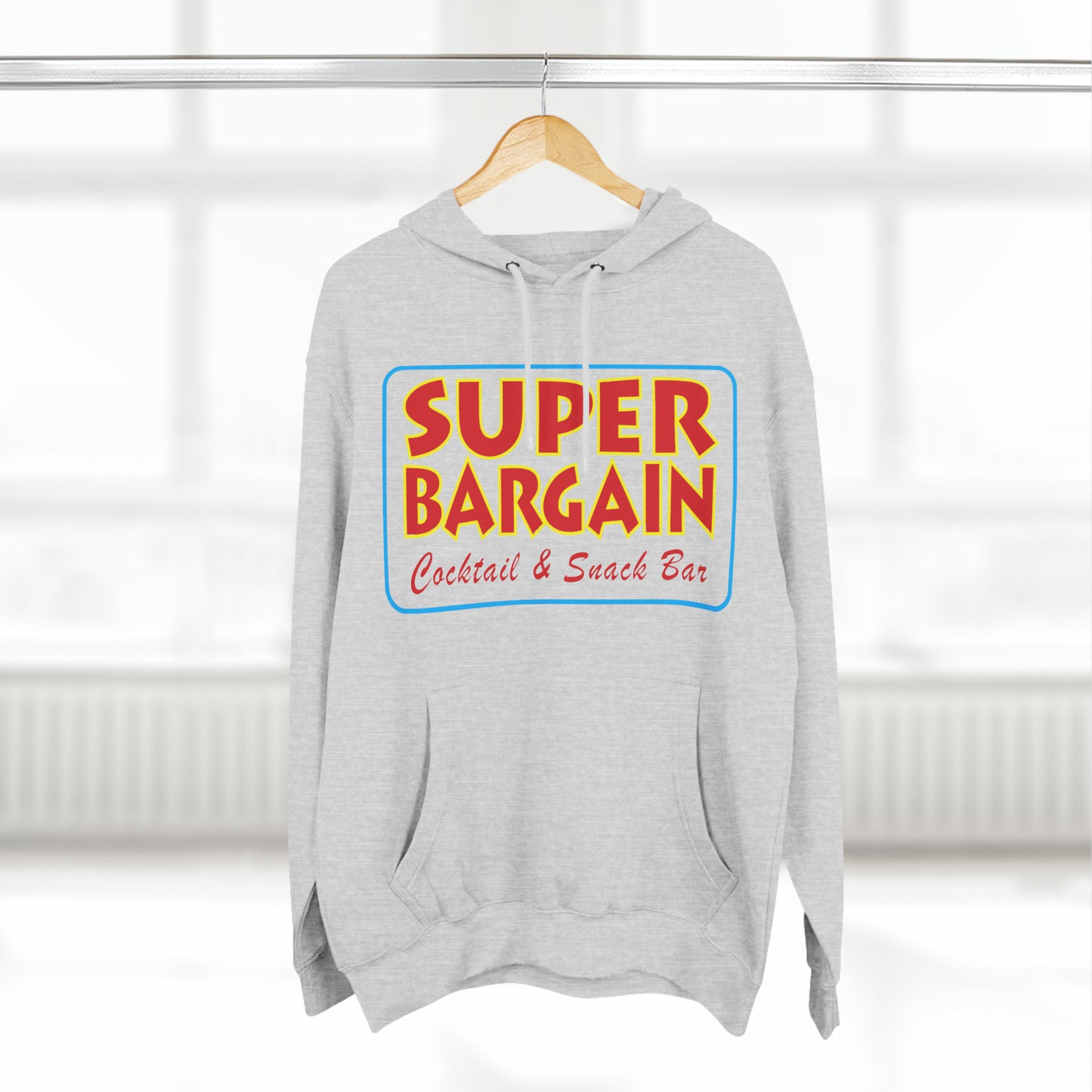 A grey Unisex Premium Pullover Hoodie with the words "SUPER BARGAIN Cocktail & Snack Bar" in bold, colorful lettering, hanging on a rack in a bright, sunlit room in Cabbagetown, Toronto.