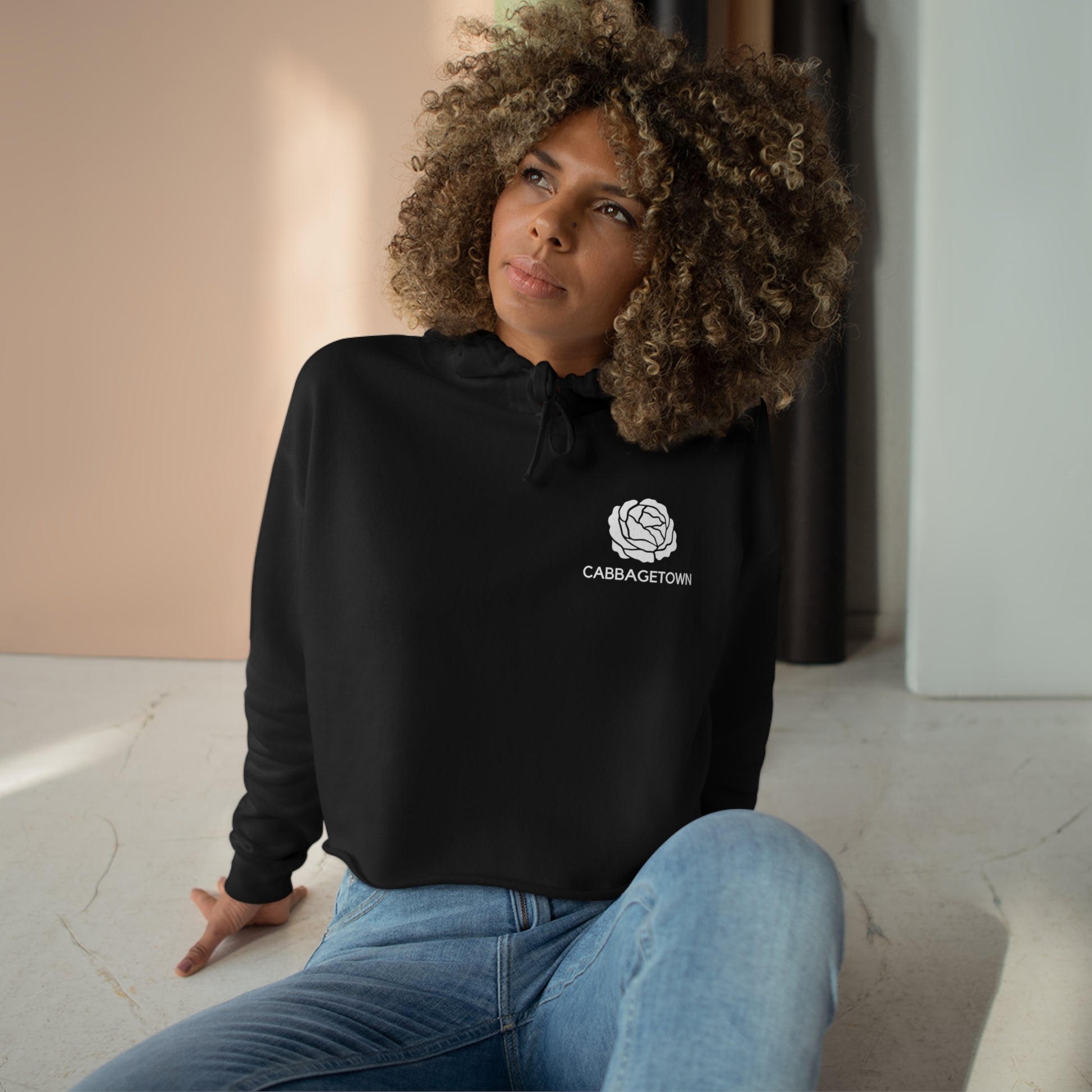 A woman with curly hair sitting on the floor, wearing jeans and a Crop Hoodie with Monochrome Cabbagetown and Super Bargain Logo by Printify, looking thoughtfully to the side.