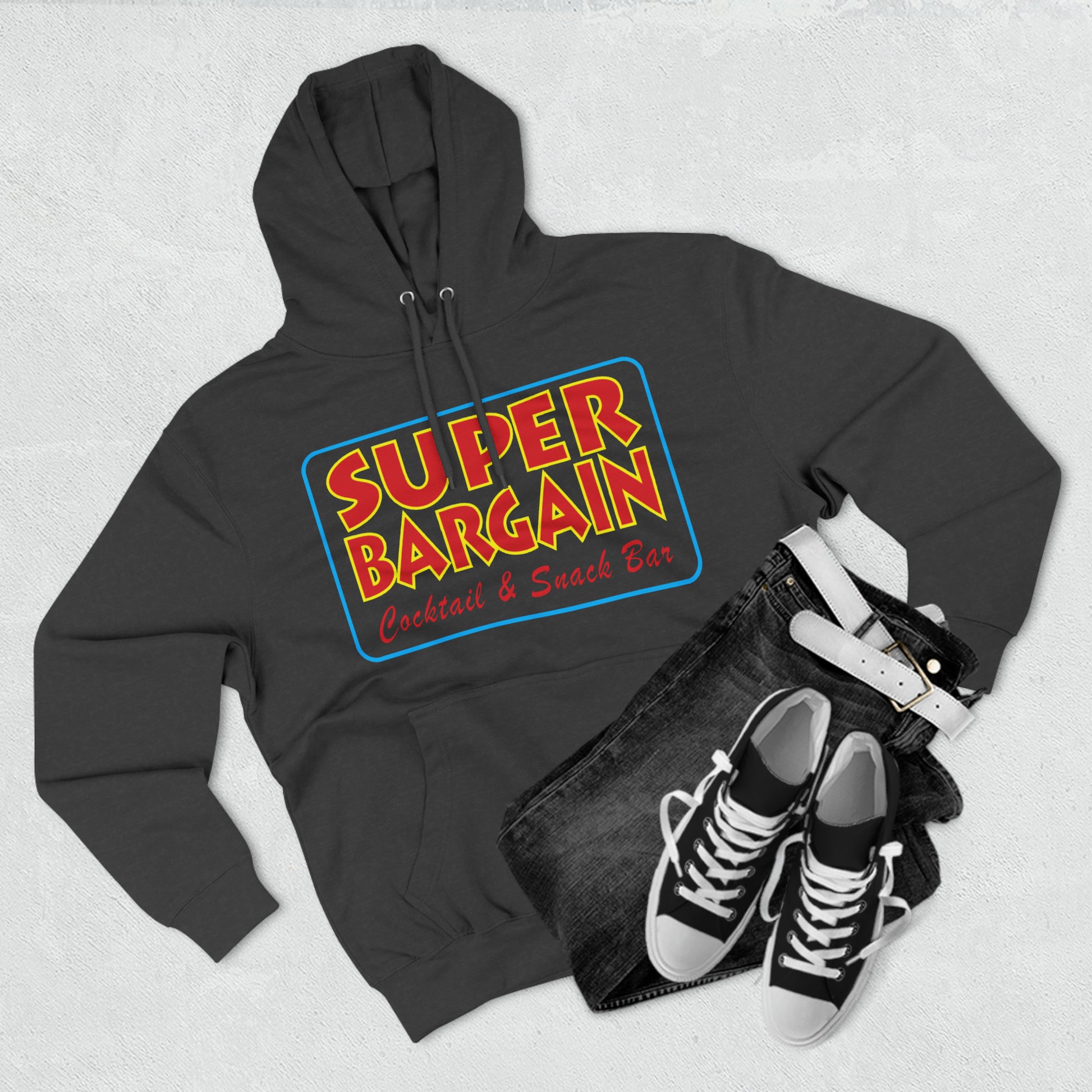 A stylish flat lay of a gray Unisex Premium Pullover Hoodie with the words "Super Bargain Cocktail & Snack Bar - Cabbagetown, Toronto" printed in vibrant colors, paired with black jeans and black and white sneakers on a textured white background. (Brand Name: Printify)