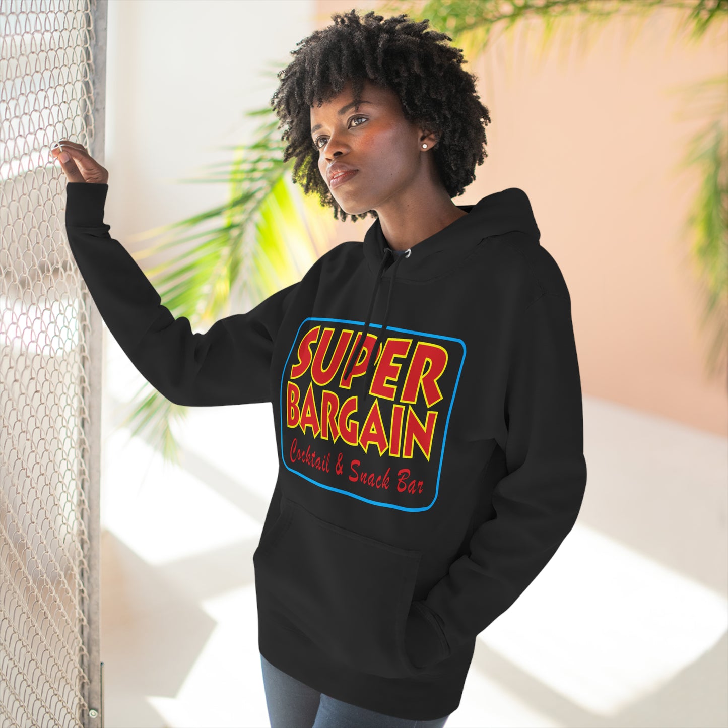A woman wearing a black Printify Unisex Premium Pullover Hoodie with the colorful text "Super Bargain Central & Snack Bar" stands by a metal fence in Cabbagetown, Toronto, looking to the side thoughtfully.