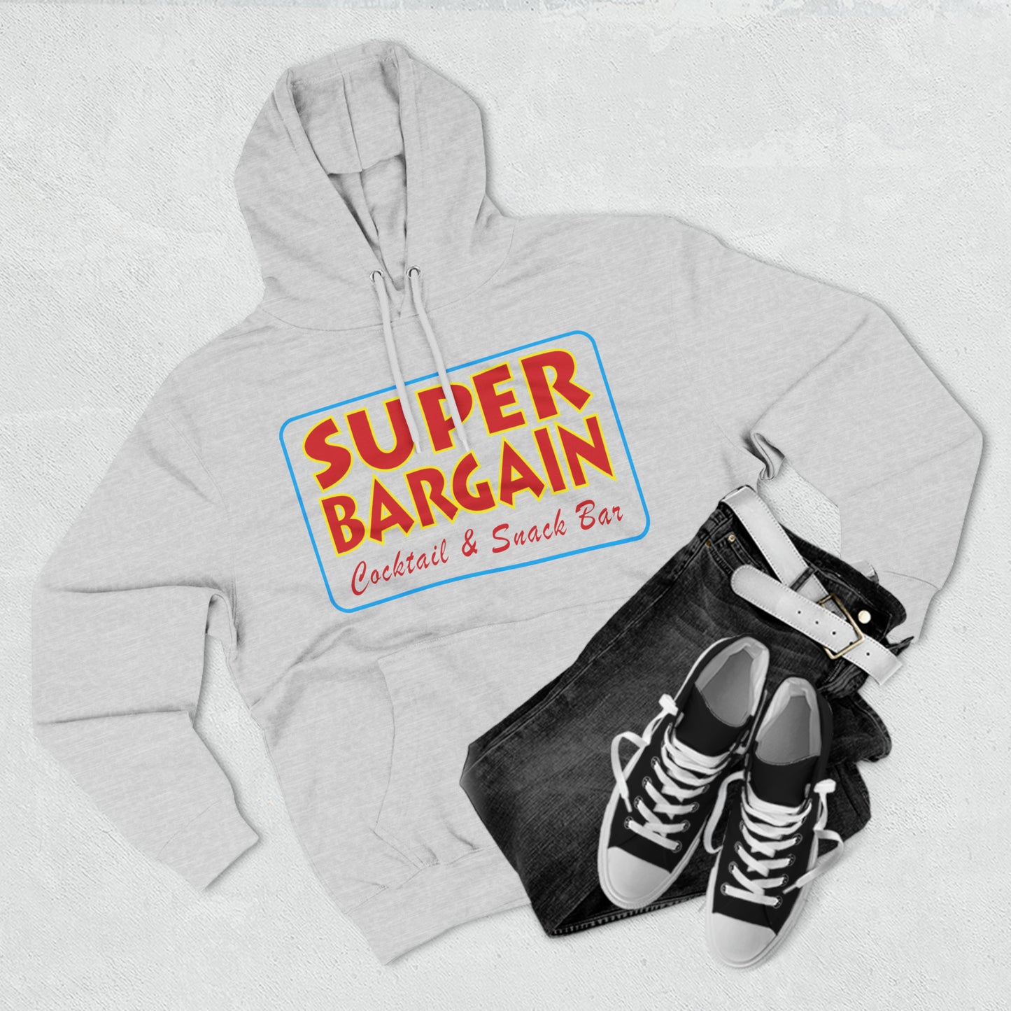 A flat lay of a gray "Printify Unisex Premium Pullover Hoodie" with "Cabbagetown Super Bargain Cocktail & Snack Bar" logo, paired with dark jeans and black sneakers, all arranged on a textured white background.
