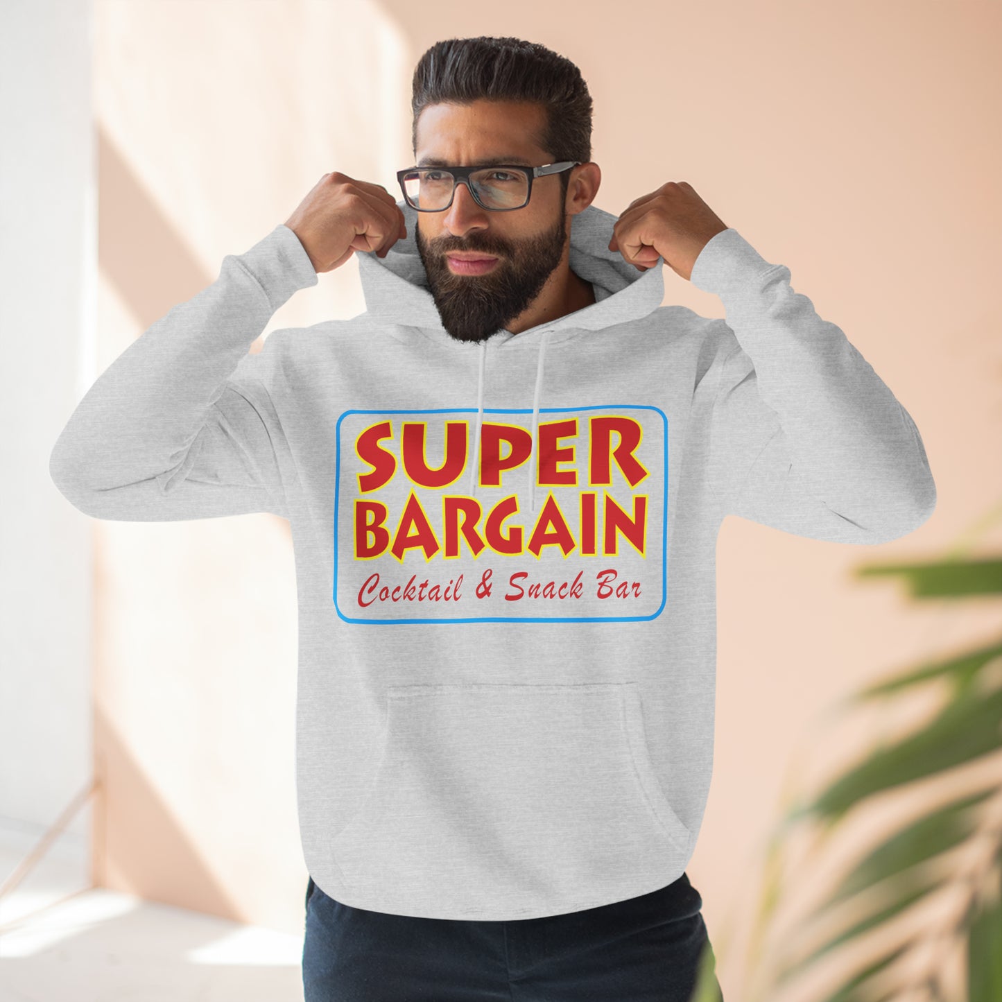 A bearded man wearing glasses adjusts the hood of his gray Printify Unisex Premium Pullover Hoodie, which features a colorful "Super Bargain Cocktail & Snack Bar" logo, popular in Cabbagetown, Toronto, on the front. Light filters softly into the room.