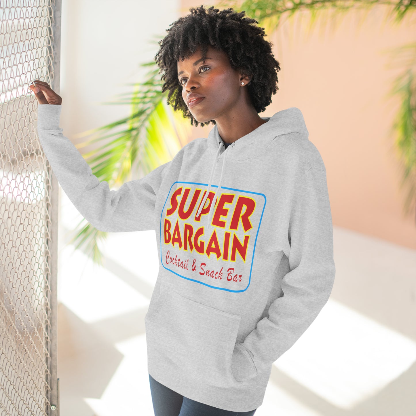 A woman with curly hair wearing a gray Printify Unisex Premium Pullover Hoodie printed with "Super Bargain Cereal & Snack Bar" stands by a metallic fence in Cabbagetown, Toronto, looking thoughtfully to the side, with tropical plants in the background.
