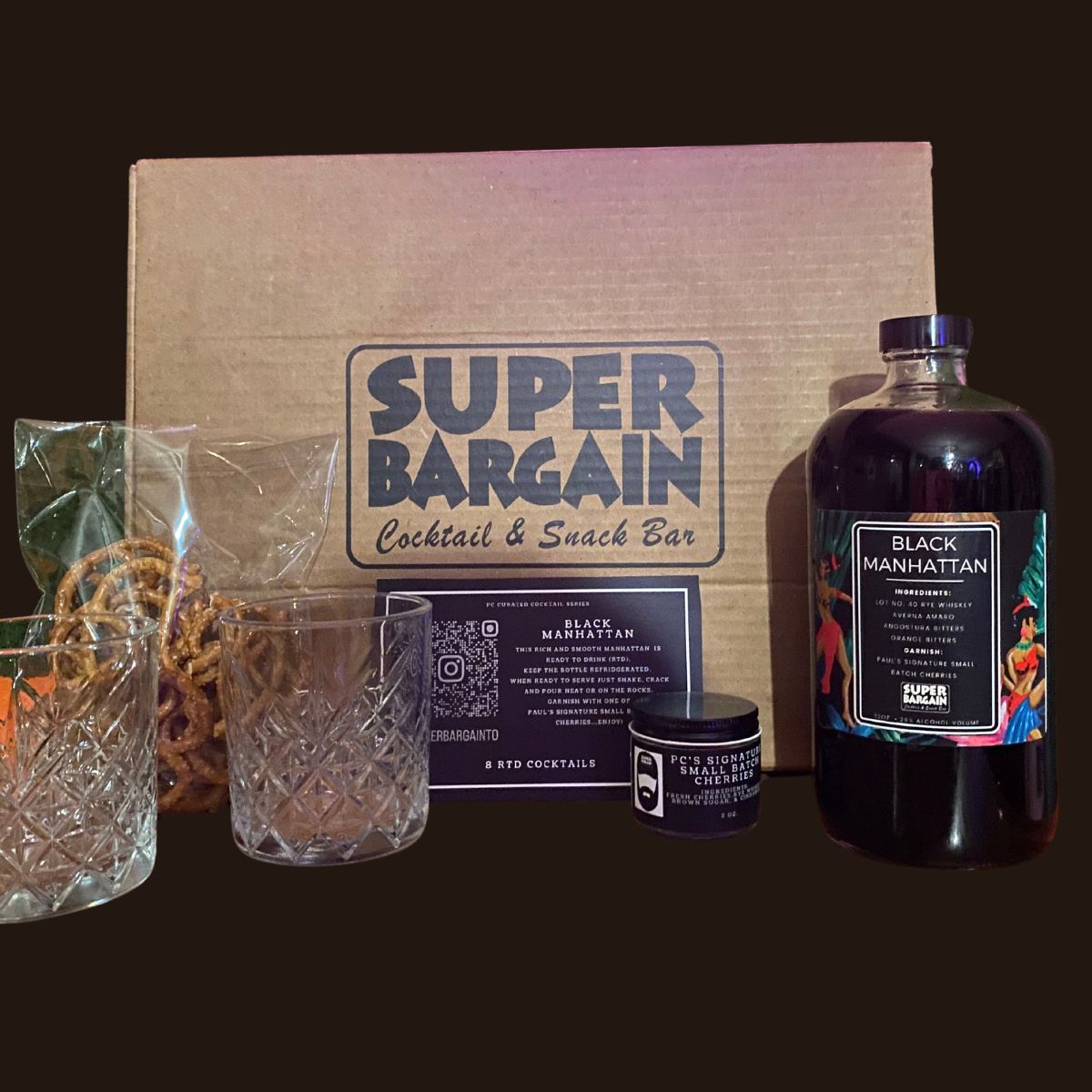 A cocktail kit labeled "Super Bargain Cocktail & Snack Bar," featuring a bottle of premium bourbon "Black Manhattan RTD Cocktail Gift Box, two crystal glasses, snacks in clear bags, and a small candle, arranged on a dark surface.