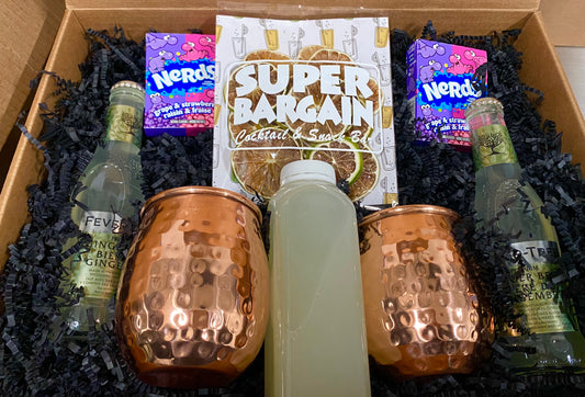 A gift box containing two copper mugs, bottles of Fever-Tree ginger ale, a bottle of lime juice, and packets of Nerds candy, arranged on black crinkle paper, with a "Pomegranate Mule RTD Cocktail Kit" sign centered above. Brand: Super Bargain Shop