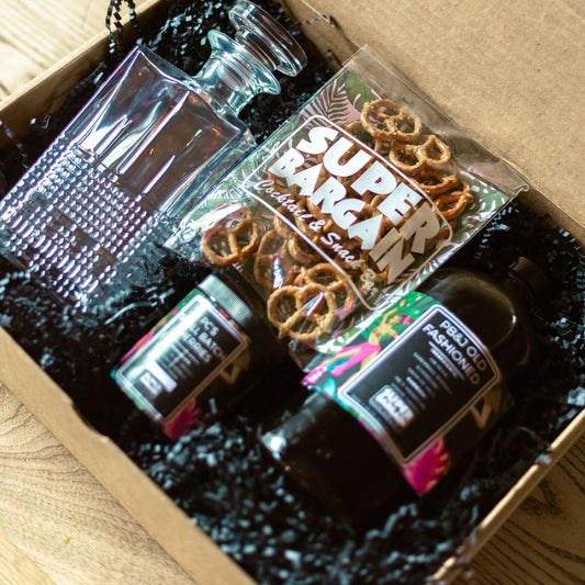 A gift box containing a bottle of Black Manhattan or PB&J Old Fashioned Premium Spirit RTD Cocktail Gift Box, a glass decanter, a small jar, and a bag of pretzels labeled "Super Bargain Shop." Black crinkle paper cushions the items within the cardboard box. The box is resting on a wooden surface.