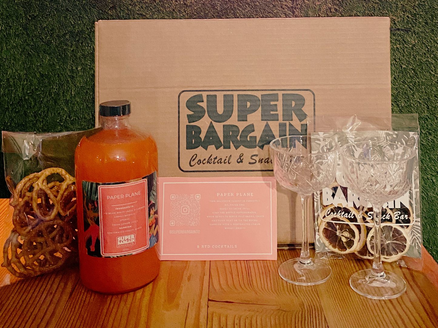 A **Paper Plane RTD Cocktail Gift Box** featuring a cardboard box labeled "**Super Bargain Shop**," a bottle of RTD Paper Plane, four empty crystal glasses, a bag of pretzels, and a bag with dried citrus slices is displayed on a wooden surface.