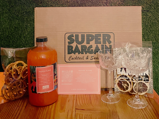 A **Paper Plane RTD Cocktail Gift Box** featuring a cardboard box labeled "**Super Bargain Shop**," a bottle of RTD Paper Plane, four empty crystal glasses, a bag of pretzels, and a bag with dried citrus slices is displayed on a wooden surface.