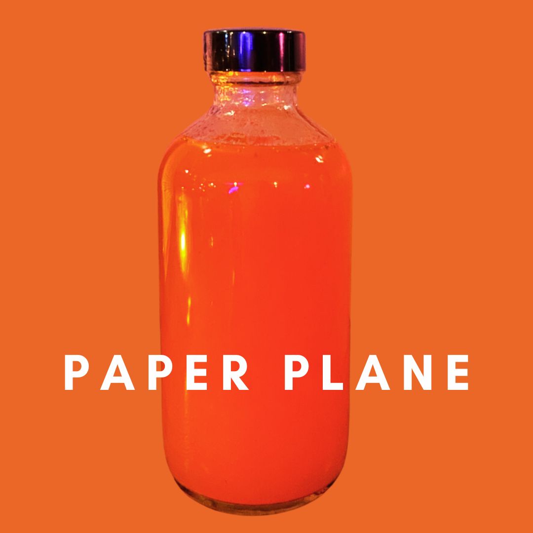 A clear glass bottle filled with a vibrant orange liquid is set against an orange background. The bottle, with a shiny black cap, could be part of an elegant gift box. The words "Paper Plane RTD Cocktail Gift Box" by Super Bargain Shop are written in bold white letters near the bottom of the image, hinting at a handcrafted cocktail experience.