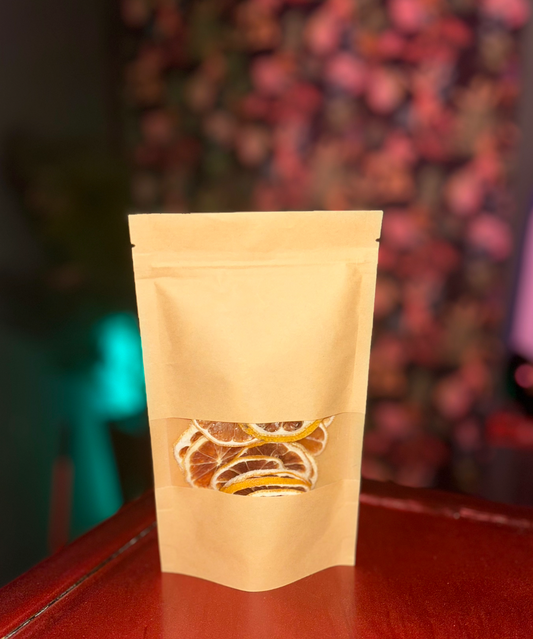 A brown paper pouch stands on a shiny red surface, with Super Bargain Shop dehydrated lemon slices visible through a clear window on the front, and a blurred floral background.