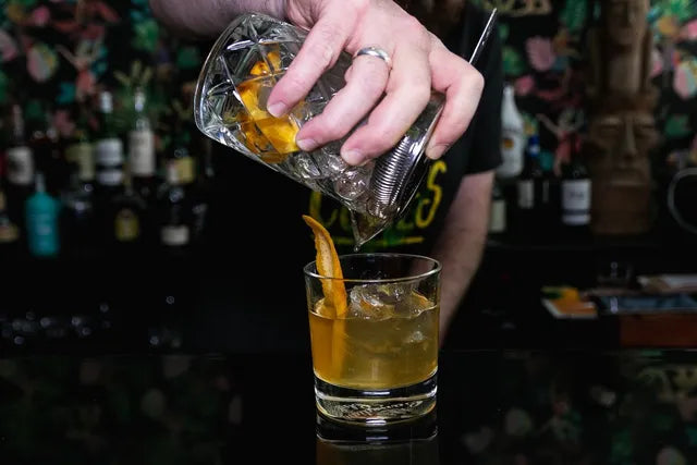 Bartender pouring a drinkg into a cocktail glass with one hand. The hand has 1 ring on it and the cocktail is light orange with a few pieces of ice and a orange swirl.